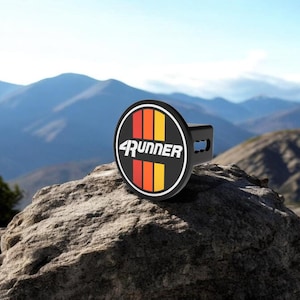 May include: A black circular hitch cover featuring the white text 4RUNNER centered over three vertical stripes in red, orange, and yellow. The hitch cover has a square mounting bracket extending from the back, designed for a vehicle receiver.