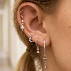 May include: A collection of silver earrings adorns an ear, featuring various celestial designs. The earrings include small studs, a crescent moon, and dangling stars, all embellished with sparkling accents. The jewelry is displayed against a blurred background.