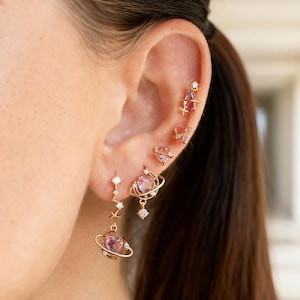 Puede incluir: Un conjunto de pendientes dorados con diseños celestiales. Los pendientes incluyen pendientes de botón con forma de planeta con gemas rosas, pendientes de estrella y planeta colgantes, y pendientes de botón en forma de corazón. Los pendientes se llevan en la oreja, mostrando los diferentes estilos.