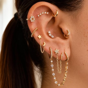 Op de afbeelding: Een oor met meerdere gouden oorbellen. De oorbellen omvatten creolen, studs en hangende ontwerpen met sterren, manen en sprankelende stenen. De sieraden zijn geplaatst tegen de achtergrond van iemands donkere haar en een vage gang.