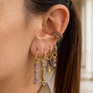 Puede incluir: Una colección de pendientes dorados con piedras preciosas de colores. Los pendientes incluyen gotas colgantes de amatista, un diseño geométrico con un triángulo y un brazalete de oreja arcoíris. Las joyas se exhiben en una oreja.