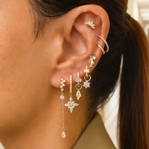 Könnte beinhalten: Ein Ohr, das mit mehreren goldenen Schmuckstücken geschmückt ist. Der Schmuck umfasst einen kleinen Ohrstecker, einen Doppelreifen und mehrere baumelnde Ohrringe mit klaren Edelsteinen. Die Ohrringe weisen verschiedene Formen auf, darunter Sterne, Tropfen und geometrische Designs.