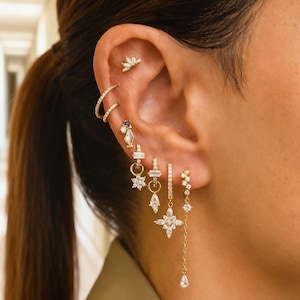 Peut inclure: Une oreille ornée de multiples boucles d'oreilles dorées. Les boucles d'oreilles comprennent un petit clou en forme de fleur, un double anneau et divers modèles pendants avec des pierres précieuses transparentes. Les bijoux sont mis en valeur sur l'oreille et les cheveux d'une personne.