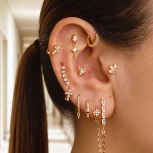 Puede incluir: Una oreja adornada con múltiples pendientes dorados de varios diseños. Estos incluyen una estrella, una cruz, una media luna y pequeños y brillantes tachuelas. Algunos pendientes presentan cadenas colgantes y formas geométricas.