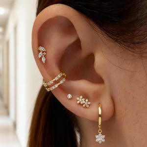 Könnte beinhalten: Nahaufnahme eines Ohrs, das mit mehreren goldenen Schmuckstücken geschmückt ist. Das Ohr hat einen kleinen Ohrstecker, einen doppelreihigen Ohrklemmen, einen Blütencluster-Ohrstecker, einen hängenden Creolen-Ohrring mit einem Blumenanhänger und einen blattförmigen Ohrstecker.