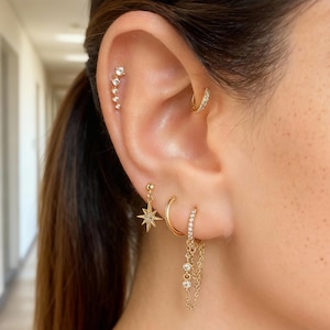 May include: An ear featuring several gold earrings. A small hoop earring, a star-shaped drop earring, and a chain earring with gems are shown. A line of small, sparkling studs runs up the ear. The jewelry is gold-toned.