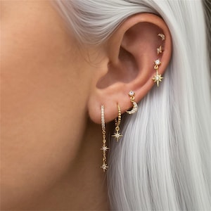 Puede incluir: Primer plano de una oreja adornada con múltiples pendientes dorados. Los pendientes incluyen una cadena colgante con tres pequeños dijes de estrella, un aro, una luna creciente con cristales incrustados y un diseño de estrella.