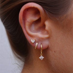Puede incluir: Primer plano de una oreja adornada con tres pendientes dorados. El pendiente superior es un pequeño pendiente en forma de lágrima. El pendiente del medio es un pequeño aro con cristales incrustados. El pendiente inferior es un aro dorado con un diseño de flor de cuatro pétalos colgante.