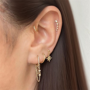 May include: Close-up of an ear with multiple gold earrings. A small hoop earring with clear stones is in the helix. A curved bar of clear stones is in the upper ear. A dangling star earring and a hoop earring with chain and clear stones are in the lobe.