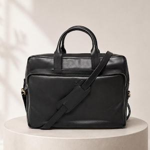 Può includere: Una borsa ventiquattrore in pelle nera con due manici superiori, uno scomparto principale con cerniera e una tracolla regolabile con una sezione imbottita. La parte anteriore presenta una tasca più piccola con cerniera e un logo impresso.