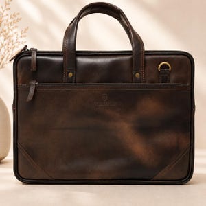 Può includere: Una ventiquattrore in pelle marrone scuro con due manici superiori, una tasca frontale con cerniera e un anello a D in ottone. La borsa ha una forma rettangolare con angoli rinforzati, cuciture a vista e un logo impresso al centro.