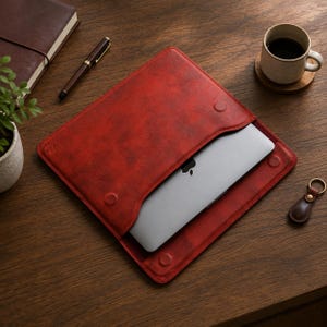 Könnte beinhalten: Eine dunkelrote, texturierte Laptoptasche mit Klappverschluss und zwei runden Magnetverschlüssen. Ein silberner Laptop mit einem schwarzen Apfel-Logo ist teilweise in die Hülle eingeschoben und zeigt das metallische Finish.