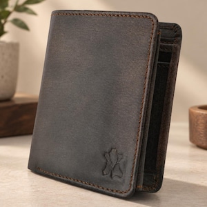 Könnte beinhalten: Ein dunkelbraunes Leder-Bifold-Portemonnaie mit sichtbaren Kontrastnähten an den Kanten und einem kleinen geprägten Logo in der unteren rechten Ecke. Das Portemonnaie steht aufrecht und zeigt eine glatte, matte Textur sowie eine strukturierte rechteckige Form.