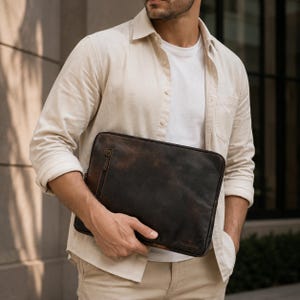 Könnte beinhalten: Eine dunkelbraune Laptoptasche aus Leder in rechteckiger Form mit einer vertikalen Reißverschlusstasche an der Seite und einem dezent geprägten Logo in der unteren rechten Ecke. Die Tasche hat eine glatte, abgenutzte Oberfläche.