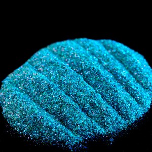 May include: A close-up of teal blue glitter with iridescent shimmer. The glitter is piled in a mound and has a textured surface.