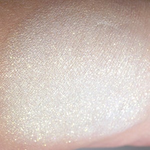 May include: Close-up of a swatch of shimmering, pearlescent makeup. The product has a light, creamy color with a subtle sparkle. The texture appears smooth and finely milled, reflecting light to create a luminous effect.