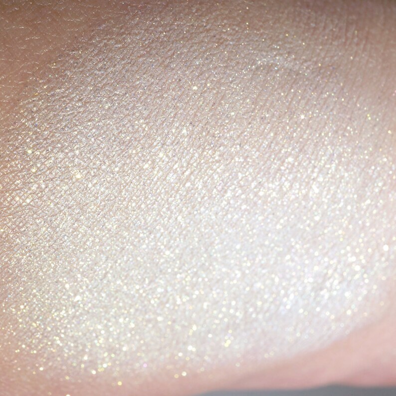 May include: Close-up of a swatch of shimmering, pearlescent makeup. The product has a light, creamy color with a subtle sparkle. The texture appears smooth and finely milled, reflecting light to create a luminous effect.