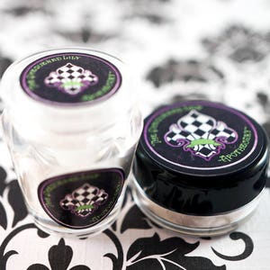 May include: Two small apothecary jars with black lids and labels. The labels feature a checkered fleur-de-lis design in black, white, and purple, with the text "The Squeezed Lily Apothecary". One jar is clear, the other is black.