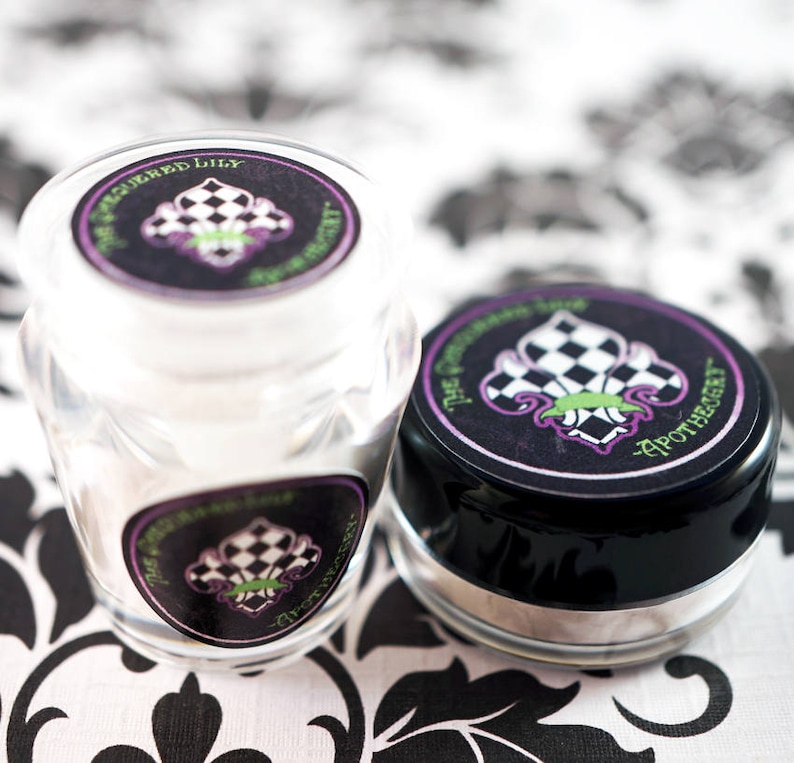 May include: Two small apothecary jars with black lids and labels. The labels feature a checkered fleur-de-lis design in black, white, and purple, with the text "The Squeezed Lily Apothecary". One jar is clear, the other is black.