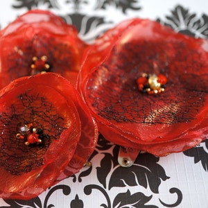Puede incluir: Tres flores de tela roja con red negra y detalles dorados. Las flores están superpuestas y tienen un aspecto delicado y femenino.