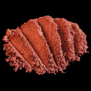 May include: A close-up of a pile of red-brown powder, likely a natural pigment or cosmetic, arranged in a wave-like pattern against a black background.