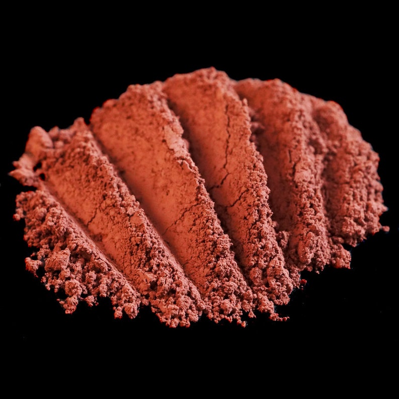 May include: A close-up of a pile of red-brown powder, likely a natural pigment or cosmetic, arranged in a wave-like pattern against a black background.