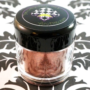 May include: A small, clear jar with a black lid containing a shimmery, copper-colored loose eyeshadow. The lid has a black and white checkered design with a gold fleur-de-lis and the text "The Gypsy  Lady Apothecary".