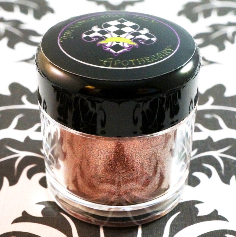 May include: A small, clear jar with a black lid containing a shimmery, copper-colored loose eyeshadow. The lid has a black and white checkered design with a gold fleur-de-lis and the text "The Gypsy  Lady Apothecary".