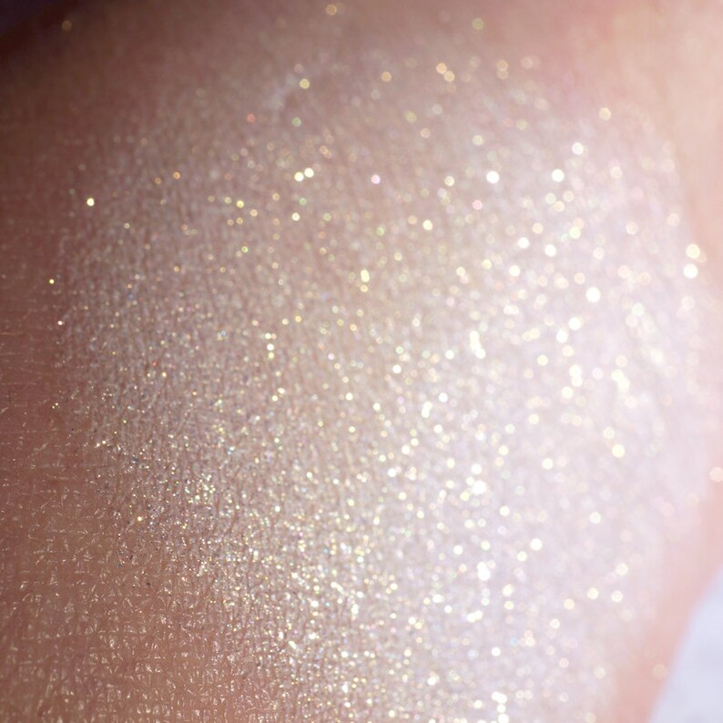 May include: Close-up of a shimmering, light-toned cosmetic product. The product has a pearlescent sheen with scattered glitter particles, creating a luminous effect. The texture appears smooth with a subtle, reflective quality.