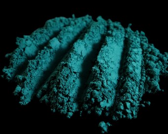 May include: Close-up of a pile of teal-colored powder with visible ridges against a black background. The powder has a fine, granular texture, and the ridges create a sense of depth and dimension. The color is vibrant and saturated.