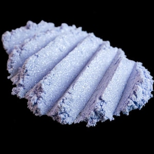 Pode incluir: Close-up de um monte de pó cosmético azul claro com um acabamento brilhante e cintilante. O pó é disposto em um padrão texturizado e canelado contra um fundo preto, destacando sua cor e textura. O produto parece ser sombra para os olhos ou um cosmético semelhante.