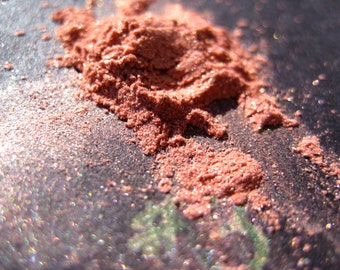 May include: Close-up of a pile of loose, shimmery, peach-colored eyeshadow powder on a dark, textured surface.