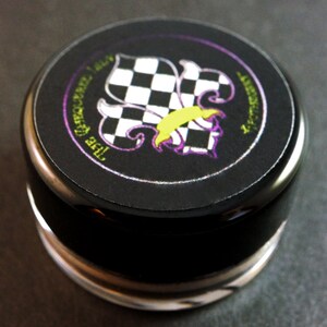 May include: Black round container with a silver lid. The lid has a black and white checkered fleur-de-lis design with purple and yellow accents. The text "The  Experience" is printed around the design.