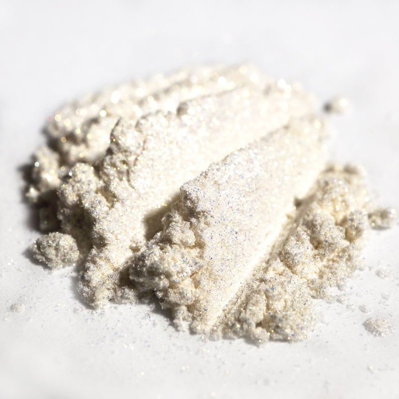 May include: Close-up of a pile of shimmering, off-white cosmetic powder. The powder has a textured, granular appearance with iridescent highlights, suggesting a pearlescent or glittery finish. The product is likely a highlighter or eyeshadow.