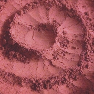 May include: Close-up of a pink powder with a textured surface. The powder is arranged in a swirling pattern, creating a heart shape in the center.