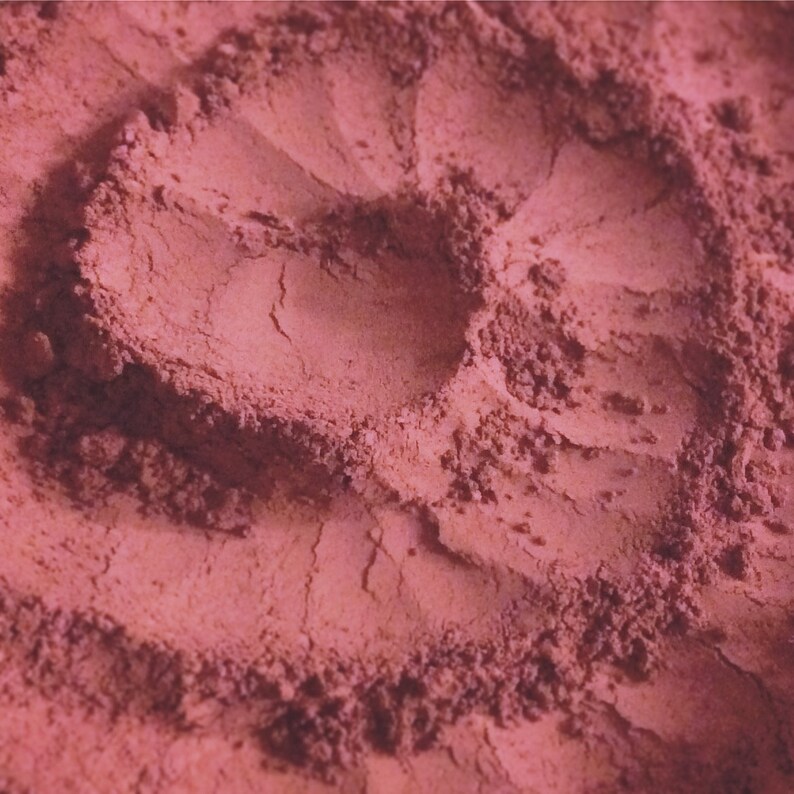 May include: Close-up of a pink powder with a textured surface. The powder is arranged in a swirling pattern, creating a heart shape in the center.