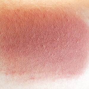 May include: A close-up of a warm, reddish-brown blush swatch on skin. The blush has a smooth, matte finish.