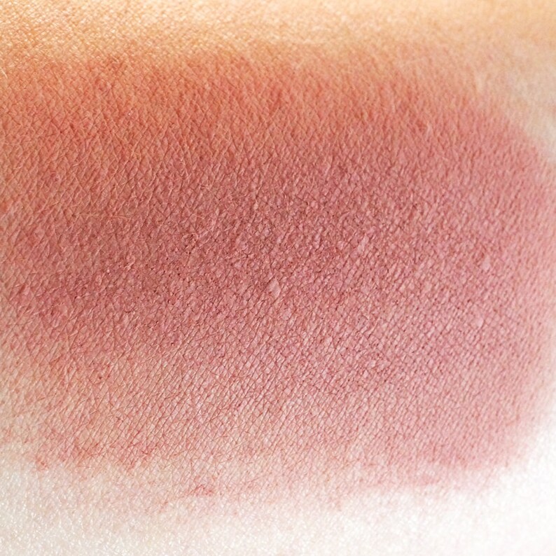 May include: A close-up of a warm, reddish-brown blush swatch on skin. The blush has a smooth, matte finish.