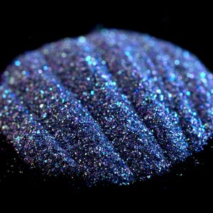 May include: A close-up of iridescent blue glitter with a slight purple hue. The glitter is piled in a mound and has a textured, sparkly appearance.