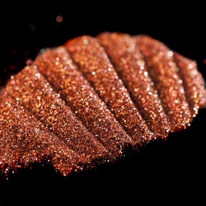May include: Close-up of a pile of copper glitter with a shimmery, iridescent finish. The glitter is arranged in a wave-like pattern, creating a textured and sparkly effect.