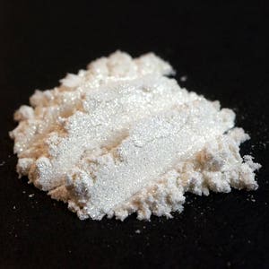May include: A pile of shimmering, white cosmetic powder against a black background. The powder has a pearlescent sheen, catching the light and appearing slightly iridescent. The texture is fine and granular, suggesting a loose powder product.