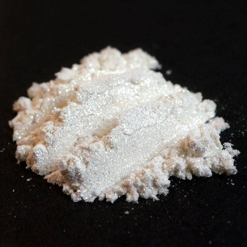 May include: A pile of shimmering, white cosmetic powder against a black background. The powder has a pearlescent sheen, catching the light and appearing slightly iridescent. The texture is fine and granular, suggesting a loose powder product.