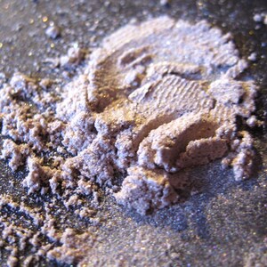 Pu&ograve; includere: Primo piano di un mucchio di glitter sciolto di colore viola chiaro con una finitura scintillante. Il glitter &egrave; sparso su una superficie grigio scuro.