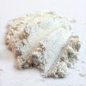 May include: A close-up of a pile of white, shimmering cosmetic powder. The powder has a pearlescent sheen and appears finely milled, with visible glitter particles. The texture is slightly granular, suggesting a loose powder formulation, ideal for makeup.