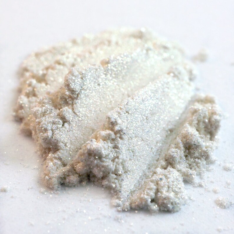 May include: A close-up of a pile of white, shimmering cosmetic powder. The powder has a pearlescent sheen and appears finely milled, with visible glitter particles. The texture is slightly granular, suggesting a loose powder formulation, ideal for makeup.