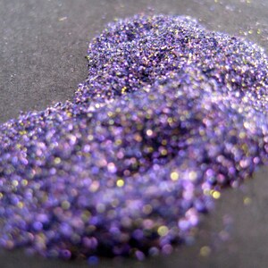 May include: Close-up of a pile of iridescent purple glitter. The glitter sparkles with hints of gold and other colors, creating a shimmering effect. The glitter is on a dark gray surface.