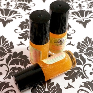 May include: Three small, cylindrical bottles with black caps and orange liquid. The bottles have labels with text and are filled with glitter. The bottles are on a white surface with a black floral pattern.