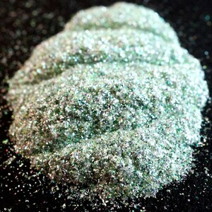 May include: A pile of iridescent green glitter with a pearlescent finish. The glitter is loose and scattered on a black surface.