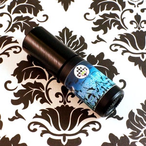 Op de afbeelding: Een zwarte rollerfles met een blauw etiket met de tekst "Blue Candy Floss". Het etiket bevat ook de woorden "Sweet, Thrt, Blue Raspberry, Aroma & Spun Sugar". De fles staat op een zwart-wit bloemenpatroon.