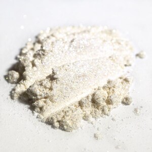 May include: Close-up of a pile of shimmering, pearlescent powder. The powder has a fine texture and reflects light, creating a sparkling effect. The color is a soft, off-white with hints of silver and gold. This product is likely a cosmetic, such as eyeshadow or highlighter.